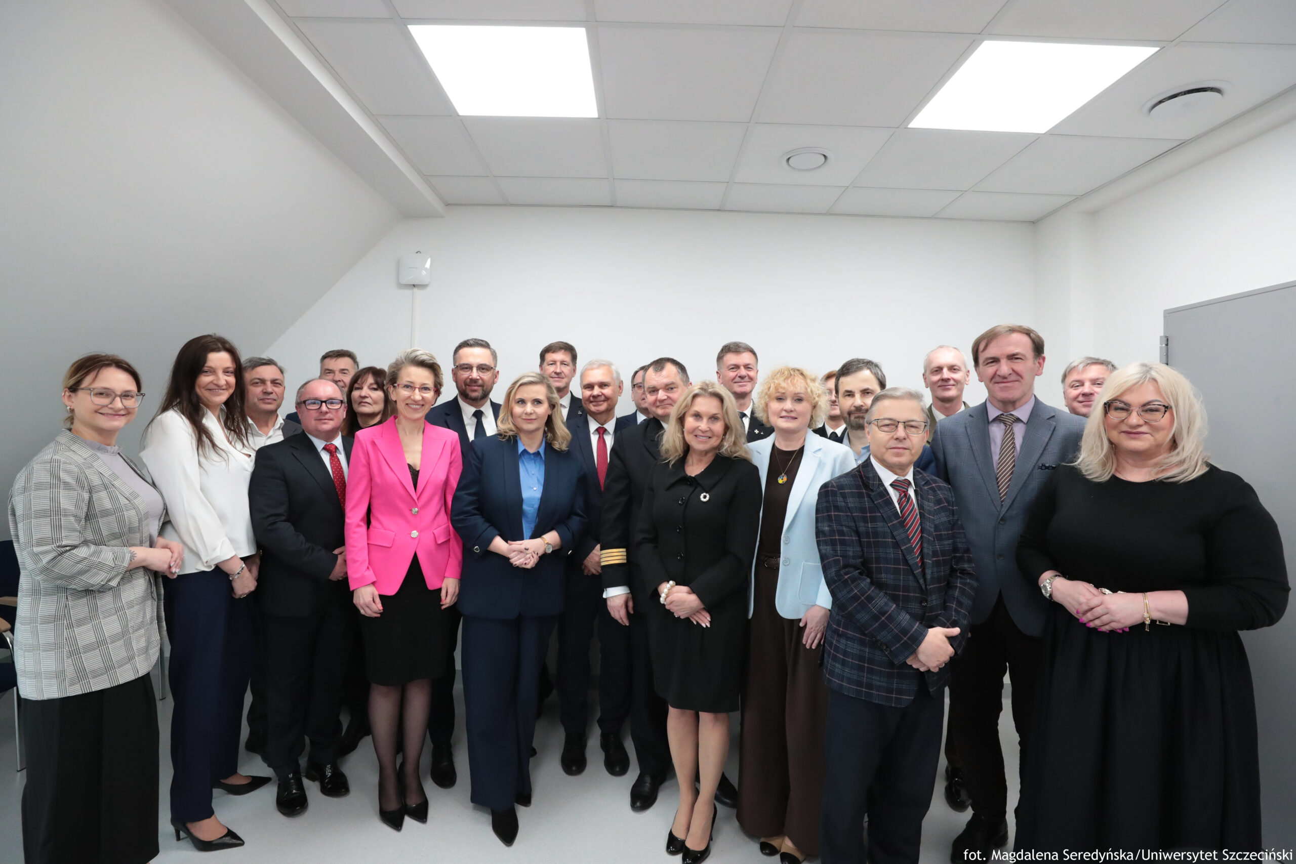Grupowe zdjęcie około dwudziestu osób stojących w kilku rzędach w jasnym, nowoczesnym pomieszczeniu. To głównie dorośli w średnim wieku; większość ma na sobie formalne stroje biznesowe. Pomieszczenie ma białe ściany i sufit z kwadratowych paneli z dużymi, prostokątnymi lampami. Po prawej stronie widać szare drzwi. Na pierwszym planie stoją kobiety i mężczyźni w garniturach i eleganckich zestawach. W centrum wyróżnia się kobieta w intensywnie różowej marynarce i czarnej spódnicy oraz kobieta w granatowym garniturze z jasną koszulą. Obok nich stoi kobieta w czarnym stroju z jasnymi włosami, a po prawej kobieta w jasnej (białej lub bardzo jasnoszarej) marynarce. Z przodu po prawej stoi mężczyzna w kraciastej marynarce z krawatem, a przy samym prawym brzegu kobieta w czarnej sukience z jasnymi, blond włosami i okularami. Na dole po prawej znajduje się podpis: „fot. Magdalena Seredyńska/Uniwersytet Szczeciński”.