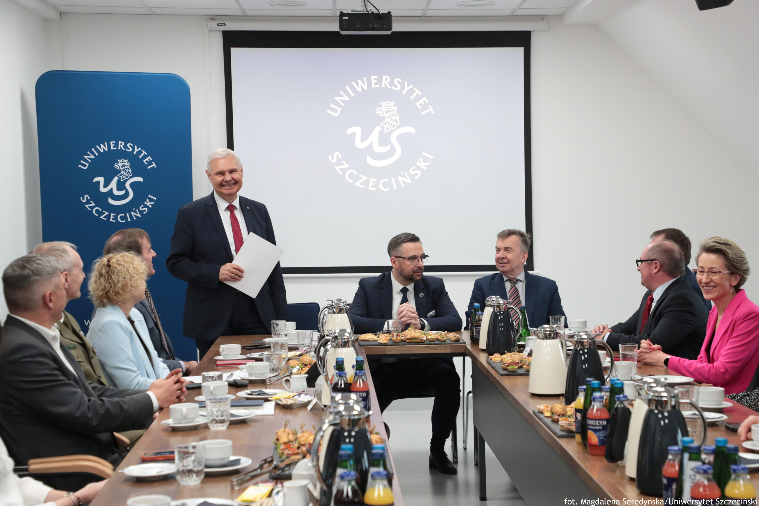 Sala konferencyjna z dwoma długimi stołami ustawionymi w kształt litery U i grupą około kilkunastu osób w strojach formalnych siedzących po obu stronach. Na środku z tyłu stoi mężczyzna w ciemnym garniturze i czerwonym krawacie, uśmiechnięty, trzymający kartkę; przed nim siedzą uczestnicy spotkania. W tle jest duży ekran projekcyjny z jasnym tłem i wyświetlonym logo oraz napisem: UNIWERSYTET SZCZECIŃSKI. Po lewej stronie stoi niebieski pionowy baner z tym samym logo i napisem: UNIWERSYTET SZCZECIŃSKI. Na stołach stoją dzbanki/termosy (czarne i białe), szklanki z wodą, filiżanki ze spodkami, talerzyki, notatniki oraz półmiski z przekąskami (małe kanapki i wypieki). Widoczne są także butelki z napojami, w tym soki i woda. W prawym dolnym rogu zdjęcia widnieje podpis: fot. Magdalena Seredyńska/Uniwersytet Szczeciński.