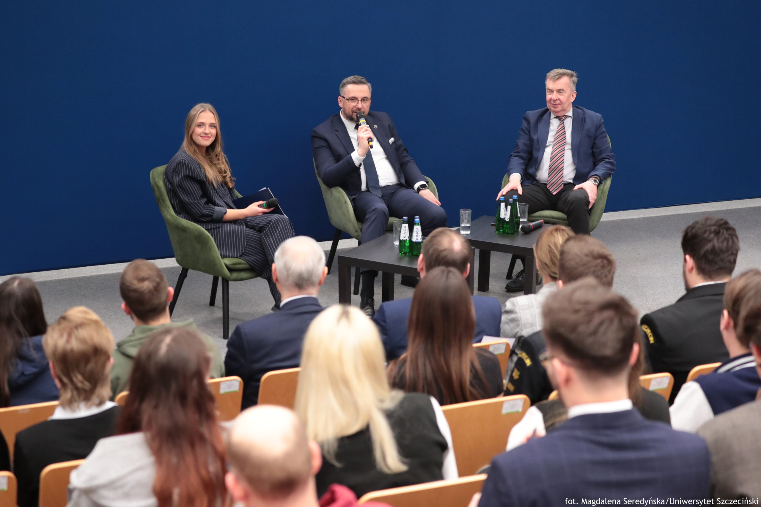 Panel dyskusyjny w sali wykładowej z trzema osobami siedzącymi w zielonych fotelach przed granatową ścianą. Na pierwszym planie widać publiczność siedzącą w rzędach. Po lewej siedzi kobieta z długimi, jasnobrązowymi włosami, w ciemnej, prążkowanej sukience; trzyma w dłoniach notatnik lub małe urządzenie. Na środku siedzi mężczyzna w ciemnym garniturze, białej koszuli i krawacie; trzyma mikrofon przy ustach, wygląda jakby mówił. Po prawej siedzi starszy mężczyzna w ciemnym garniturze i koszuli z krawatem w ukośne paski; ręce opiera na podłokietnikach. Między fotelami stoją dwa niskie, czarne stoliki. Na stolikach są zielone butelki wody mineralnej, szklanki i leżący mikrofon. W prawym dolnym rogu jest mały napis: „fot. Magdalena Seredyńska/Uniwersytet Szczeciński”.