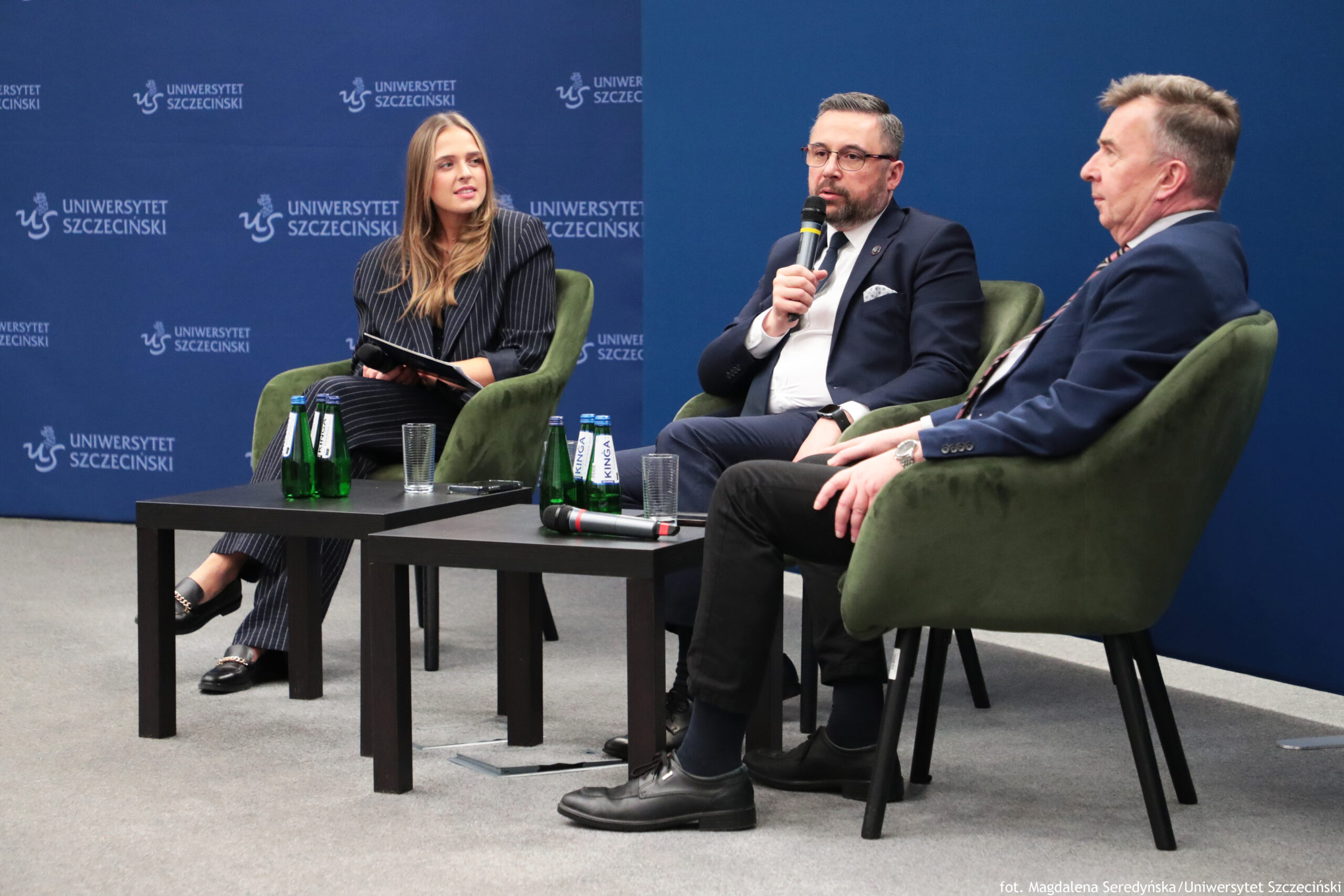 Panel dyskusyjny z trzema osobami siedzącymi w zielonych, tapicerowanych fotelach na scenie. W tle znajduje się granatowa ścianka z powtarzającymi się napisami „UNIWERSYTET SZCZECIŃSKI” oraz logo „US”. Po lewej siedzi kobieta o długich, jasnobrązowych włosach, w ciemnym garniturze w cienkie jasne prążki. Trzyma na kolanach notatnik lub teczkę i jest zwrócona w stronę dwóch pozostałych osób. Na środku siedzi mężczyzna w ciemnym garniturze, białej koszuli i krawacie, w okularach, z krótką brodą i wąsami. Trzyma mikrofon przy ustach, jak podczas wypowiedzi. Po prawej siedzi mężczyzna w granatowej marynarce i jasnej koszuli, w profilu, zwrócony w stronę osoby mówiącej. Ma krótkie włosy. Przed fotelami stoją dwa czarne, niskie stoliki. Na nich widać kilka zielonych butelek wody, kilka przezroczystych szklanek oraz dwa mikrofony leżące na blacie. Podłoga jest szara, jak wykładzina. W prawym dolnym rogu jest mały podpis: „fot. Magdalena Seredyńska/Uniwersytet Szczeciński”.