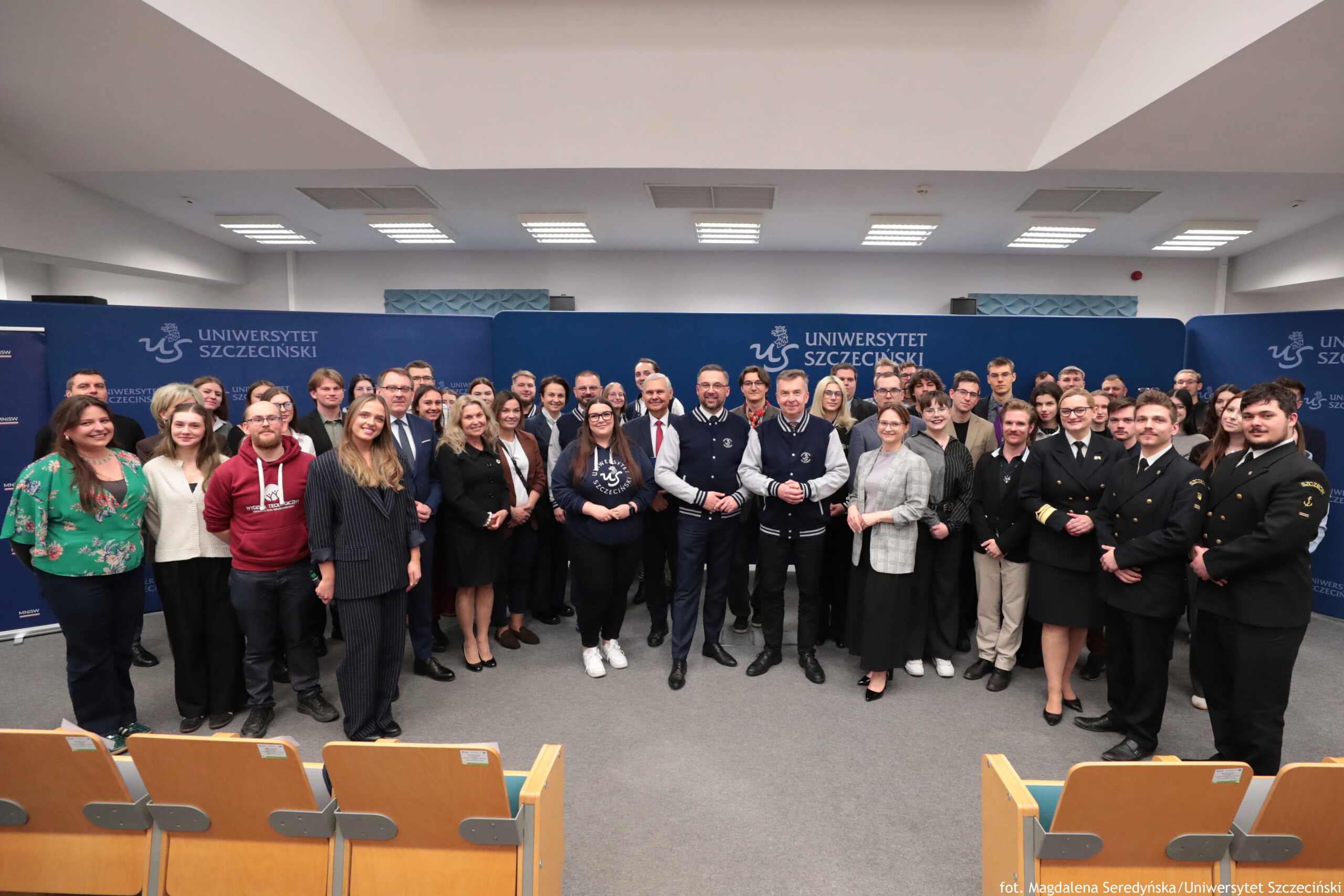 Grupowe zdjęcie kilkudziesięciu osób stojących w sali wykładowej na tle granatowej ścianki z napisem „UNIWERSYTET SZCZECIŃSKI” i logo „us”. Osoby ustawione są w kilku rzędach, pozują do wspólnej fotografii, większość patrzy w stronę aparatu i ma neutralne lub lekkie uśmiechy. Wśród nich są kobiety i mężczyźni, głównie młodzi dorośli i kilka osób w średnim wieku; stroje są mieszane: od codziennych (bluzy, swetry, jeansy) po formalne (garnitury, marynarki, sukienki). Po prawej stronie stoi kilka osób w ciemnych mundurach z widocznymi dystynkcjami na rękawach. Na środku pierwszego rzędu stoją dwaj mężczyźni w granatowych kurtkach typu varsity z jasnymi rękawami; obok nich widać osoby w garniturach i eleganckich strojach. Na dole kadru są fragmenty rzędów jasnodrewnianych siedzeń z zielonym obiciem, typowych dla auli. Oświetlenie jest jasne, sufit ma prostokątne panele świetlne. W prawym dolnym rogu znajduje się podpis: „fot. Magdalena Seredyńska/Uniwersytet Szczeciński”.