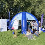 Stojący na trawie granatowo-biały, półokrągły, dmuchany namiot z logami Uniwersytetu Szczecińskiego. Przed nim siedzący na leżakach ludzie i plakat na sztaludze z napisem: Zapraszamy na kawę z Uniwersytetem Szczecińskim. W tle drzewa.