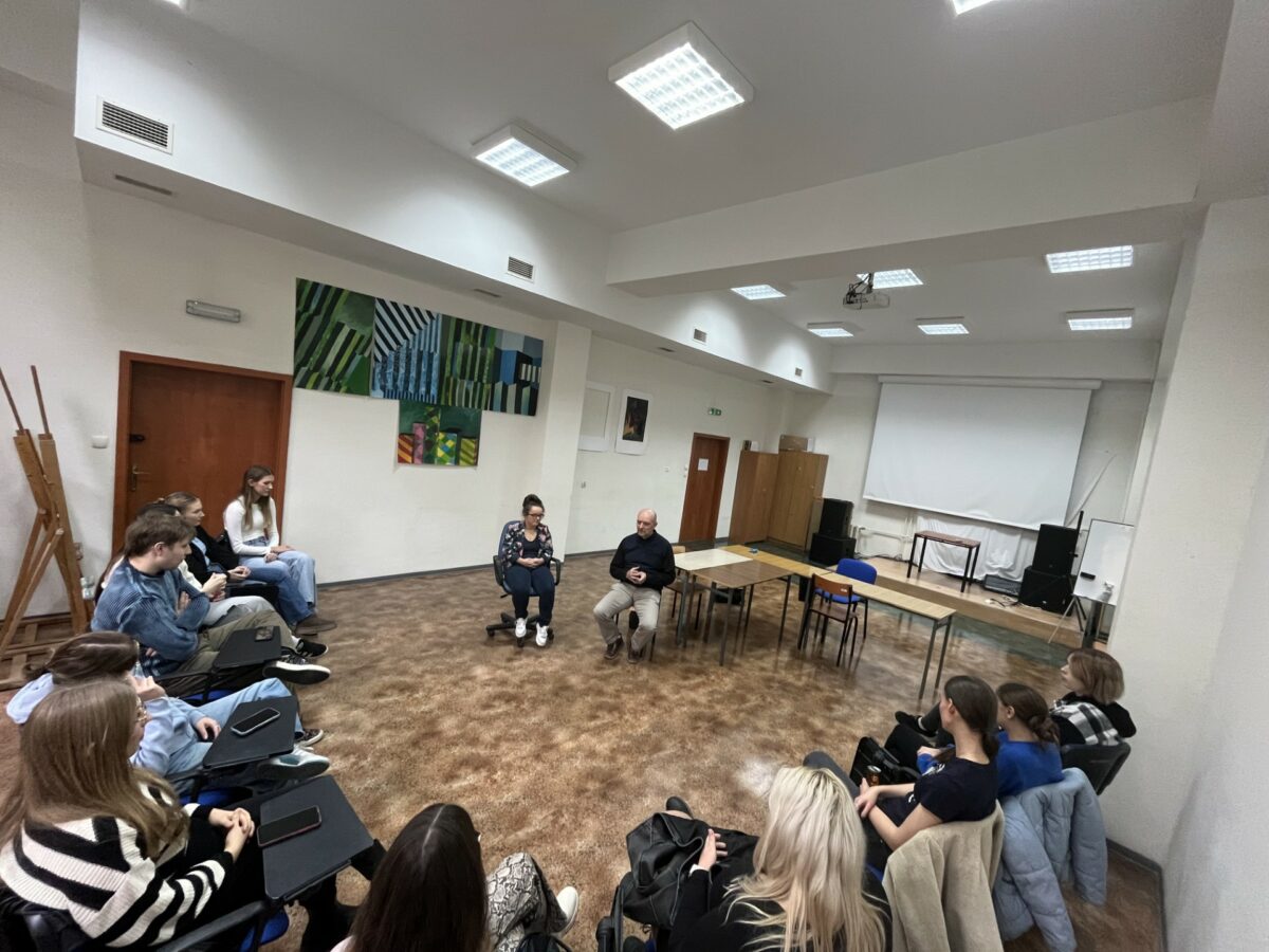 Sala wykładowa. W środku na krzesłach siedzą mężczyzna i kobieta. Dookoła nich na krzesłach siedzą studenci. W tle na jednej ze ścian wiszą obrazy w odcieniach zielonego i niebieskiego. Na ścianie po prawej stronie jest biały ekran.