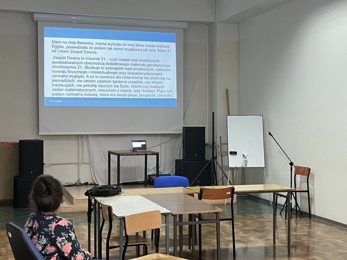 Sala wykładowa. Na pierwszym planie tyłem prelegentka na krześle (ujęcie do pasa) Obok ławki ustawione pod kątem prostym. Na ekranie prezentacja – długi opis na temat prelegentki.