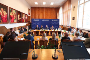 Ujęcie przodem. Ustawione w prostokąt drewniane stoły. Na pierwszym planie czarne ekrany i mikrofony. Dalej przy stołach osoby siedzące tyłem. Na kolejnym planie przodem m.in. czworo prorektorów. Po bokach pozostali uczestnicy spotkania. Na stołach szklane butelki z wodą, szklanki i filiżanki oraz dzbanki z herbatą i kawą. W tle trzy granatowe ścianki z białymi logami Uniwersytetu Szczecińskiego i logiem 40-lecia US na środkowej. Po lewej na ścianie duże portrety – poczet rektorów Uniwersytetu Szczecińskiego ubranych w gronostaje.