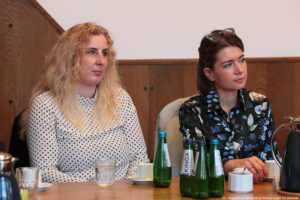 Ujęcie bokiem. Dwie kobiety siedzące przy drewnianym stole, na którym stoją szklane butelki z wodą oraz szklanki i filiżanki.