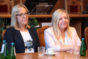 Ujęcie bokiem. Dwie kobiety siedzące przy drewnianym stole, na którym stoją szklane butelki z wodą oraz szklanki i filiżanki.