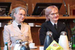 Ujęcie bokiem. Dwie kobiety siedzące przy drewnianym stole, na którym stoją szklane butelki z wodą, szklanki i filiżanki oraz dzbanki z herbatą i kawą