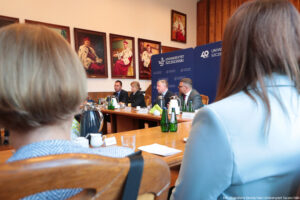 Ujęcie bokiem. Na pierwszym planie dwie osoby siedzące tyłem. Na drugim planie czworo prorektorów siedzi przy drewnianym stole przed podpisaniem porozumienia. Na stole szklane butelki z wodą, szklanki i filiżanki oraz dzbanki z herbatą i kawą. Za prorektorami granatowe ścianki z białymi logami Uniwersytetu Szczecińskiego logiem 40-lecia US. Na ścianie na wprost duże portrety – poczet rektorów Uniwersytetu Szczecińskiego ubranych w gronostaje.