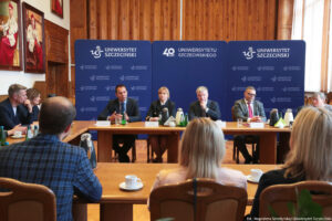 Ujęcie przodem. Na pierwszym planie trzy osoby siedzące tyłem. Na drugim planie czworo prorektorów siedzi przy drewnianym stole przed podpisaniem porozumienia. Na stole szklane butelki z wodą, szklanki i filiżanki oraz dzbanki z herbatą i kawą. W tle trzy granatowe ścianki z białymi logami Uniwersytetu Szczecińskiego i logiem 40-lecia US na środkowej.