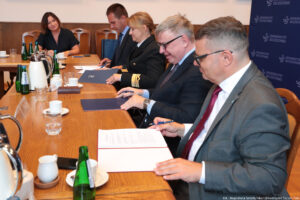 Ujęcie bokiem. Przy drewnianym stole czworo prorektorów podpisuje porozumienie. Na końcu rzeczniczka Uniwersytetu Szczecińskiego. Na stole szklane butelki z wodą, szklanki i filiżanki oraz dzbanki z herbatą i kawą.