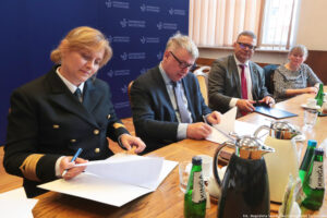 Ujęcie bokiem. Przy drewnianym stole troje prorektorów podpisuje porozumienie. Obok (po prawej) siedzi pełnomocniczka rektora Uniwersytetu Szczecińskiego. Na stole szklane butelki z wodą, szklanki i filiżanki oraz dzbanki z herbatą i kawą. W tle granatowe ścianki z białymi logami Uniwersytetu Szczecińskiego i okna z pionowymi roletami.