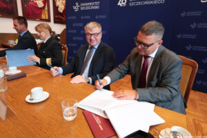 Ujęcie bokiem. Przy drewnianym stole czworo prorektorów podpisuje porozumienie. Na stole szklanki i filiżanki. W tle granatowe ścianki z białymi logami Uniwersytetu Szczecińskiego.