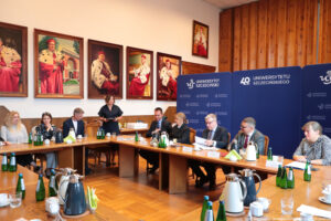 Ujęcie bokiem. Drewniane stoły ustawione pod kątem prostym. Po prawej siedzi czworo prorektorów, na końcu stojąca rzeczniczka prasowa Uniwersytetu Szczecińskiego prowadząca spotkanie. Po lewej troje przedstawicieli Biblioteki Głównej US. Na stole szklane butelki z wodą, szklanki i filiżanki oraz dzbanki z herbatą i kawą.