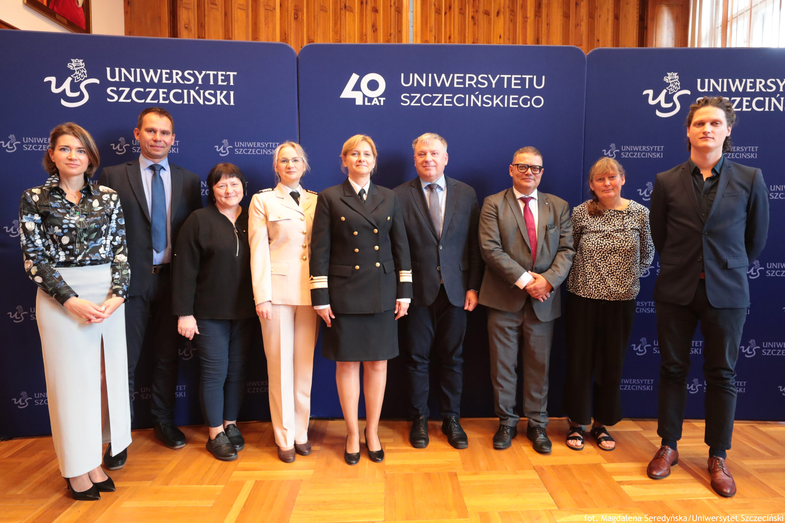 9-osobowa grupa ludzi (5 kobiet, 4 mężczyzn) stojących na tle trzech granatowych ścianek z białymi logami Uniwersytetu Szczecińskiego i logiem 40-lecia US na środkowej.