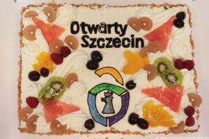 Prostokątny tort z bitą śmietaną, owocami i napisem Otwarty Szczecin oraz logiem inicjatywy w postaci otwartej kłódki z gryfem.