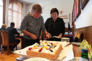 Dwie kobiety krojące prostokątny tort z bitą śmietaną, owocami i napisem Otwarty Szczecin. Na stole talerzyki, łyżeczki i chusteczki.