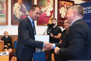 Ściskający sobie dłonie mężczyźni: prorektor Uniwersytetu Szczecińskiego z prawej strony – bokiem do kadru i z lewej prorektor Zachodniopomorskiego Uniwersytetu Technologicznego przodem do kadru. W tle mężczyzna za kamerą na statywie. Na ścianie duże portrety – poczet rektorów Uniwersytetu Szczecińskiego ubranych w gronostaje.