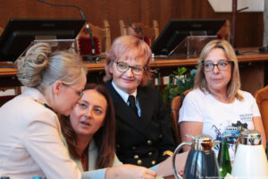 Cztery kobiety siedzące i rozmawiające przy stole (trzy po prawej przodem do kadru, jedna po lewej – bokiem). Druga kobieta od lewej trzyma kartkę, kucając. Na stole stoją dzbanki z kawą i herbatą oraz szklana butelka z wodą.