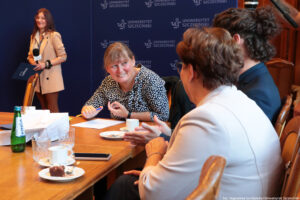 Przy drewnianych stołach ustawionych pod kątem prostym siedzą trzy kobiety (jedna przodem do kadru, dwie tyłem). Na stołach szklana butelka z wodą, szklanki i filiżanki. W tle granatowa ścianka z niebieskimi logami Uniwersytetu Szczecińskiego i dziennikarka Uniwizji – Telewizji Uniwersytetu Szczecińskiego trzymająca mikrofon.