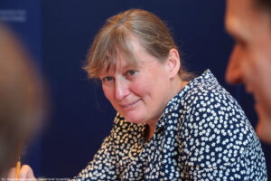 Bliski kadr. Siedząca bokiem uśmiechnięta pełnomocniczka rektora Uniwersytetu Szczecińskiego. Granatowe tło.