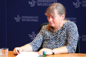 Siedząca przy drewnianym stole pełnomocniczka rektora Uniwersytetu Szczecińskiego przegląda dokumenty. Na stole szklanka z wodą. W tle granatowa ścianka z białymi logami Uniwersytetu Szczecińskiego.