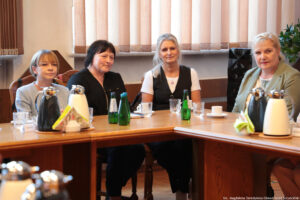 Ujęcie przodem: przy drewnianych stołach ustawionych pod kątem prostym siedzą cztery kobiety Na stołach szklane butelki z wodą, szklanki i filiżanki oraz dzbanki z herbatą i kawą. W tle okna z pionowymi roletami.