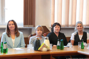 Ujęcie przodem: przy drewnianym stole siedzą cztery kobiety. Na stole szklane butelki z wodą, szklanki i filiżanki oraz dzbanki z herbatą i kawą. W tle okna z pionowymi roletami.