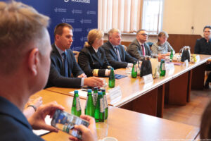 Ujęcie bokiem: przy drewnianym stole siedzi czworo prorektorów, na końcu pełnomocniczka rektora Uniwersytetu Szczecińskiego. Na stole szklane butelki z wodą, szklanki i filiżanki oraz dzbanki z herbatą i kawą. W tle okna i granatowa ścianka z niebieskimi logami US.