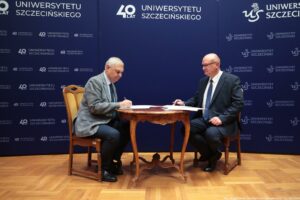 Dwóch mężczyzn w garniturach siedzących przy ozdobnym, okrągłym drewnianym stoliku i podpisujących dokumenty. Mężczyzna po lewej ma siwe włosy, jasną skórę i szarą marynarkę w kratę; trzyma długopis nad kartką. Mężczyzna po prawej jest łysy, ma jasną skórę, okulary, ciemny garnitur i krawat; także ma długopis i dokument przed sobą. Tło stanowi granatowa ścianka z powtarzającymi się napisami i logotypami „UNIWERSYTET SZCZECIŃSKI” oraz „40 LAT”. Na dole widać drewnianą podłogę. W prawym dolnym rogu jest podpis: „fot. Magdalena Seredyńska/Uniwersytet Szczeciński”.