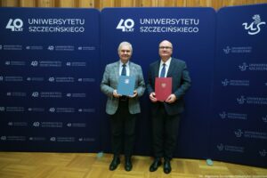 Dwóch mężczyzn w garniturach stojących obok siebie na tle granatowej ścianki z logo Uniwersytetu Szczecińskiego i napisem „40 LAT UNIWERSYTETU SZCZECIŃSKIEGO”. Mężczyzna po lewej ma siwe włosy, jasną karnację, szarą marynarkę w kratę, białą koszulę i ciemny krawat; trzyma granatową teczkę z białym okrągłym znakiem. Mężczyzna po prawej jest łysy, ma jasną karnację, ciemny garnitur, białą koszulę i ciemny krawat; trzyma czerwoną teczkę. W prawym dolnym rogu widoczny podpis: „fot. Magdalena Seredyńska/Uniwersytet Szczeciński”.