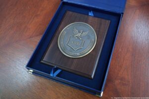 Otwarta granatowa szkatułka z drewnianą plakietą i okrągłym metalowym medalionem z godłem. Na medalionie widoczny jest orzeł nad tarczą z napisem „ABW”, a dookoła tekst: „RZECZPOSPOLITA POLSKA” oraz „AGENCJA BEZPIECZEŃSTWA WEWNĘTRZNEGO”. Szkatułka leży na drewnianym blacie. W prawym dolnym rogu jest mały podpis: „fot. Magdalena Seredyńska/Uniwersytet Szczeciński”.