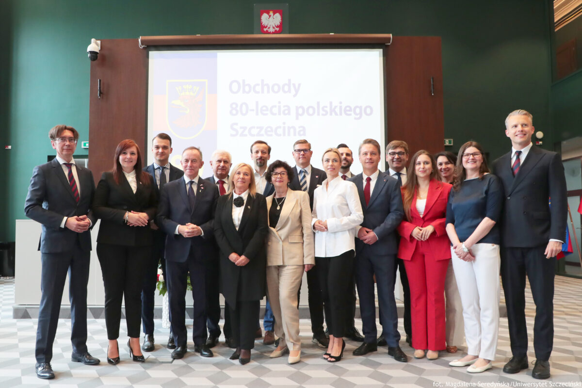 Na pierwszym planie 17 przedstawicieli Urzędu Miasta w Szczecinie. W tle na ekranie na białym tle po prawej stronie granatowy napis: „Obchody 80-lecia polskiego Szczecina”. Na ekranie po lewej szczeciński herb z czerwonym gryfem w żółtej koronie, umieszczony na tle w poziome granatowo-czerwone pasy.
