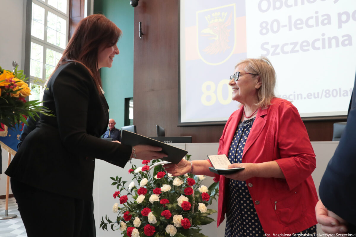 Prof. dr hab. Maria Czerepaniak-Walczak z Uniwersytetu Szczecińskiego odbierająca Medal za Zasługi dla Miasta Szczecina. Na dole stojące na podłodze biało-czerwone goździki umieszczone w białym koszu. W tle na ekranie na białym tle po prawej stronie granatowy napis: „Obchody 80-lecia polskiego Szczecina”. Na ekranie po lewej szczeciński herb z czerwonym gryfem w żółtej koronie, umieszczony na tle w poziome granatowo-czerwone pasy.