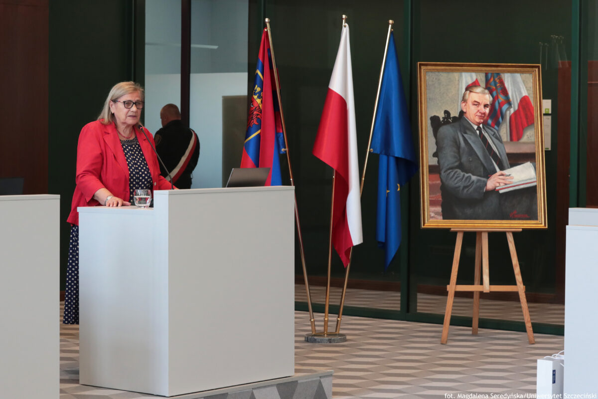 Po lewej stronie za białą mównicą z mikrofonem prof. dr hab. Maria Czerepaniak-Walczak z Uniwersytetu Szczecińskiego. Obok po prawej stronie trzy flagi na drzewcach – od lewej: granatowo-czerwona ze szczecińskim herbem, w środku polska – biało-czerwona, następnie granatowa flaga Unii Europejskiej.