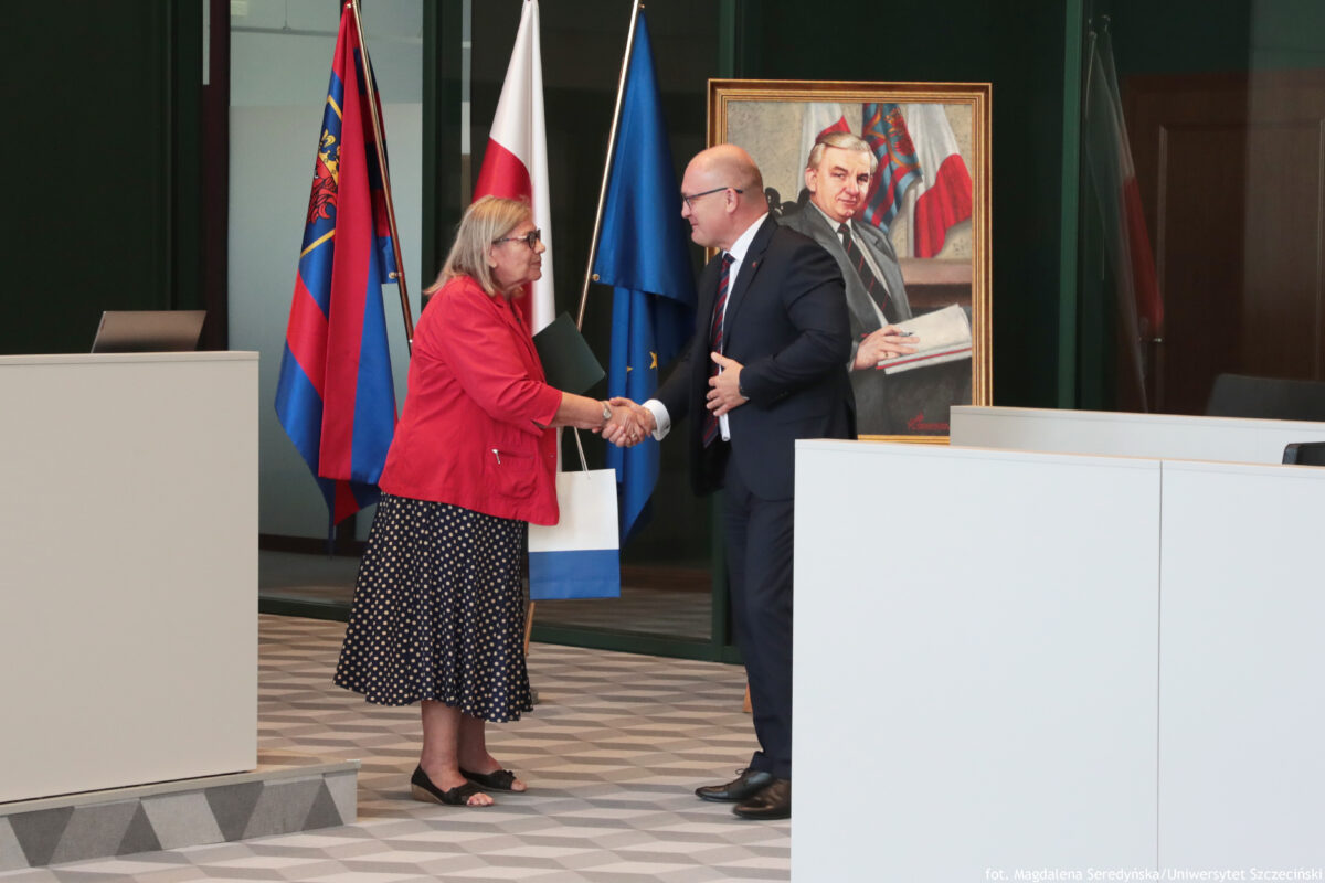 Po lewej stronie prof. dr hab. Maria Czerepaniak-Walczak z Uniwersytetu Szczecińskiego przyjmuje gratulacje od stojącego po prawej stronie Piotra Krzysta – prezydenta miasta Szczecina. W tle trzy flagi na drzewcach – od lewej: polska – biało-czerwona, w środku granatowo-czerwona ze szczecińskim herbem, następnie granatowa flaga Unii Europejskiej.
