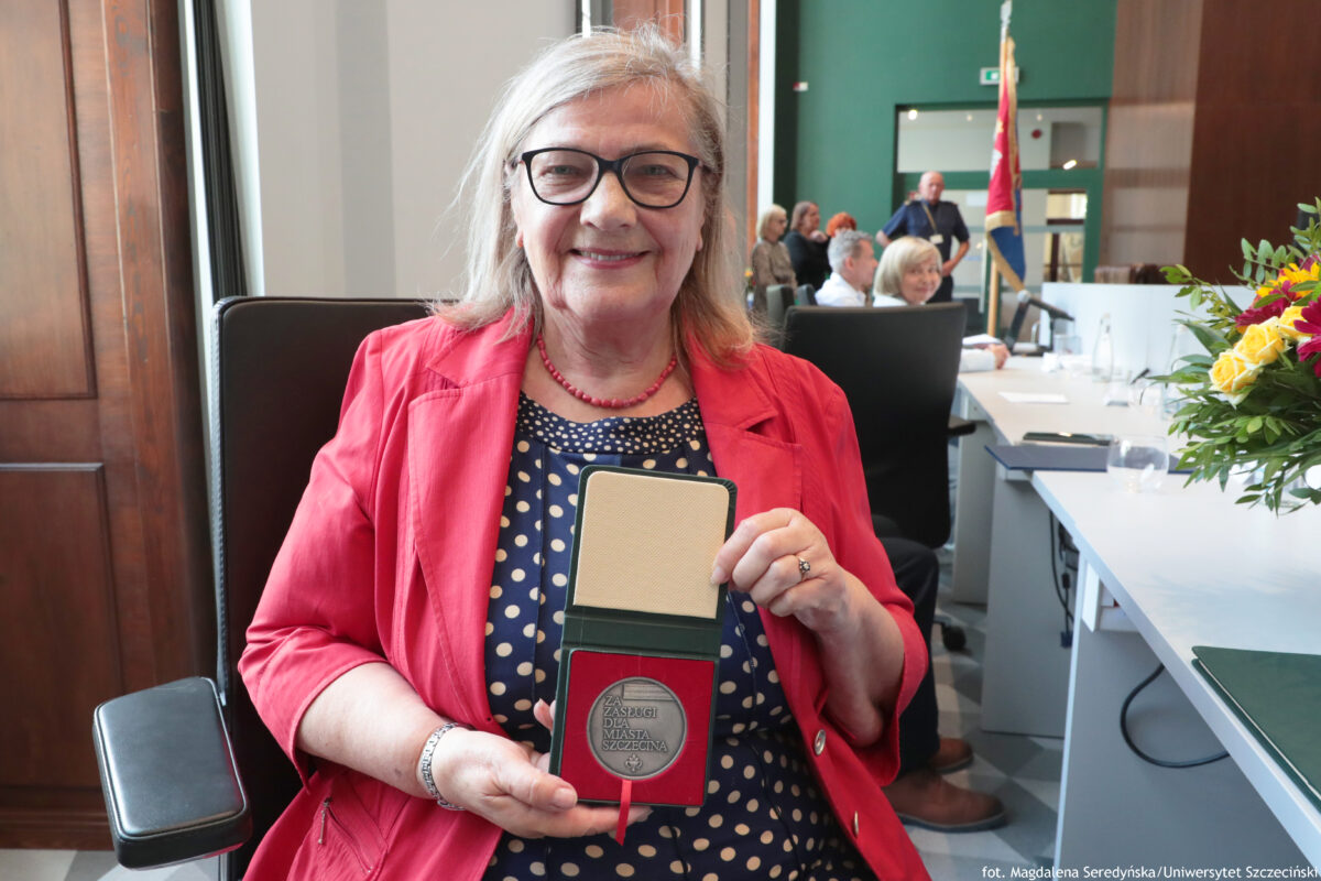 Na pierwszym planie siedzi uśmiechnięta prof. dr hab. Maria Czerepaniak-Walczak z Uniwersytetu Szczecińskiego prezentująca Medal za Zasługi dla Miasta Szczecina. Po prawej stronie na białym stole stoi bukiet kwiatów. Na końcu stołu znajduje się czerwono-granatowy sztandar.