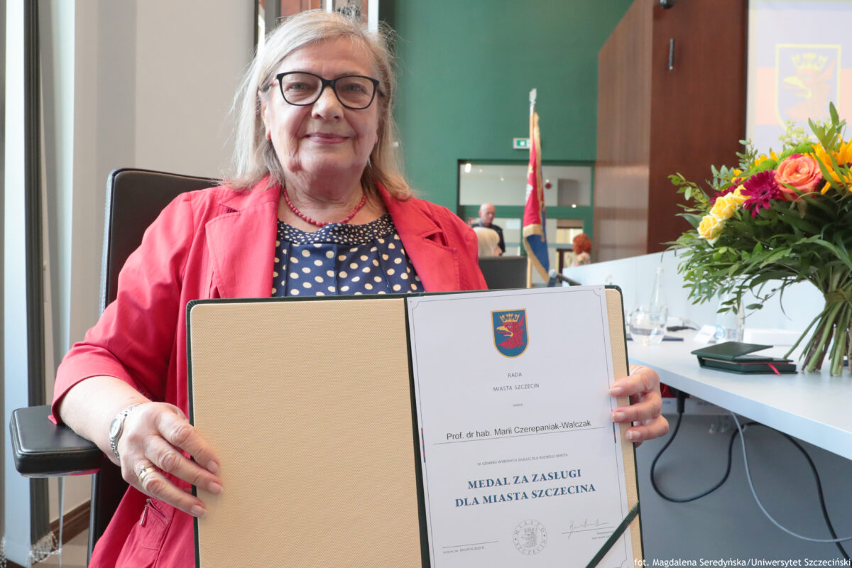 Na pierwszym planie z lewej strony uśmiechnięta prof. dr hab. Maria Czerepaniak-Walczak z Uniwersytetu Szczecińskiego prezentująca list gratulacyjny znajdujący się w eleganckiej teczce. Po prawej stronie na białym stole stoi bukiet kwiatów. Na końcu stołu znajduje się czerwono-granatowy sztandar.