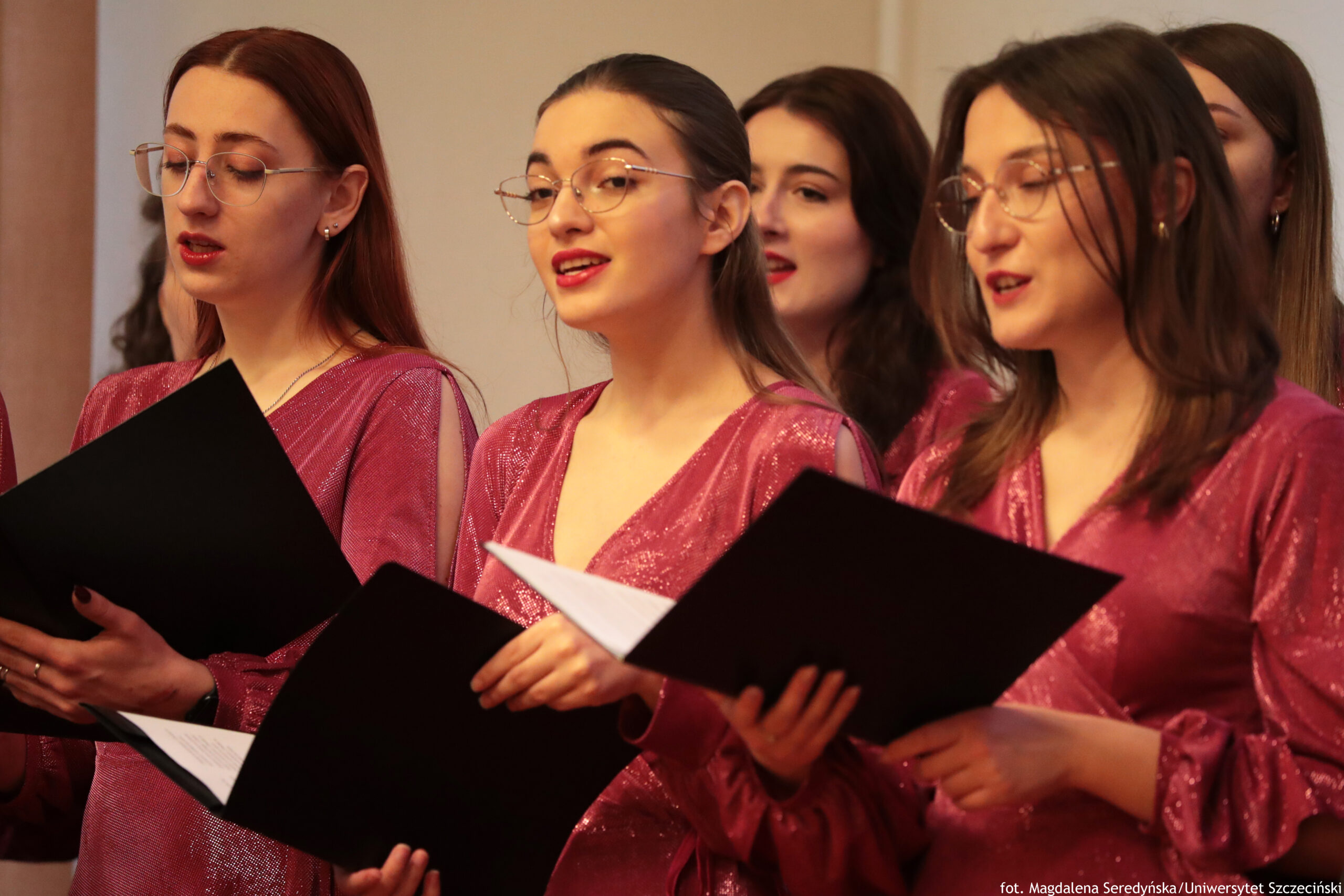 Grupa młodych kobiet śpiewających, trzymających czarne teczki z nutami. Widać pięć osób ustawionych w dwóch rzędach, w półzbliżeniu. Trzy kobiety na pierwszym planie mają jasną cerę i długie włosy; dwie z nich mają okulary. Wszystkie są ubrane w jednakowe, błyszczące sukienki w kolorze ciemnoróżowym/bordowym z dekoltem w serek i długimi rękawami. Usta kilku osób są lekko otwarte, jak podczas śpiewu. Tło jest jasne i niewyraźne, wygląda na wnętrze pomieszczenia. W prawym dolnym rogu znajduje się mały podpis: „fot. Magdalena Seredyńska/Uniwersytet Szczeciński”.