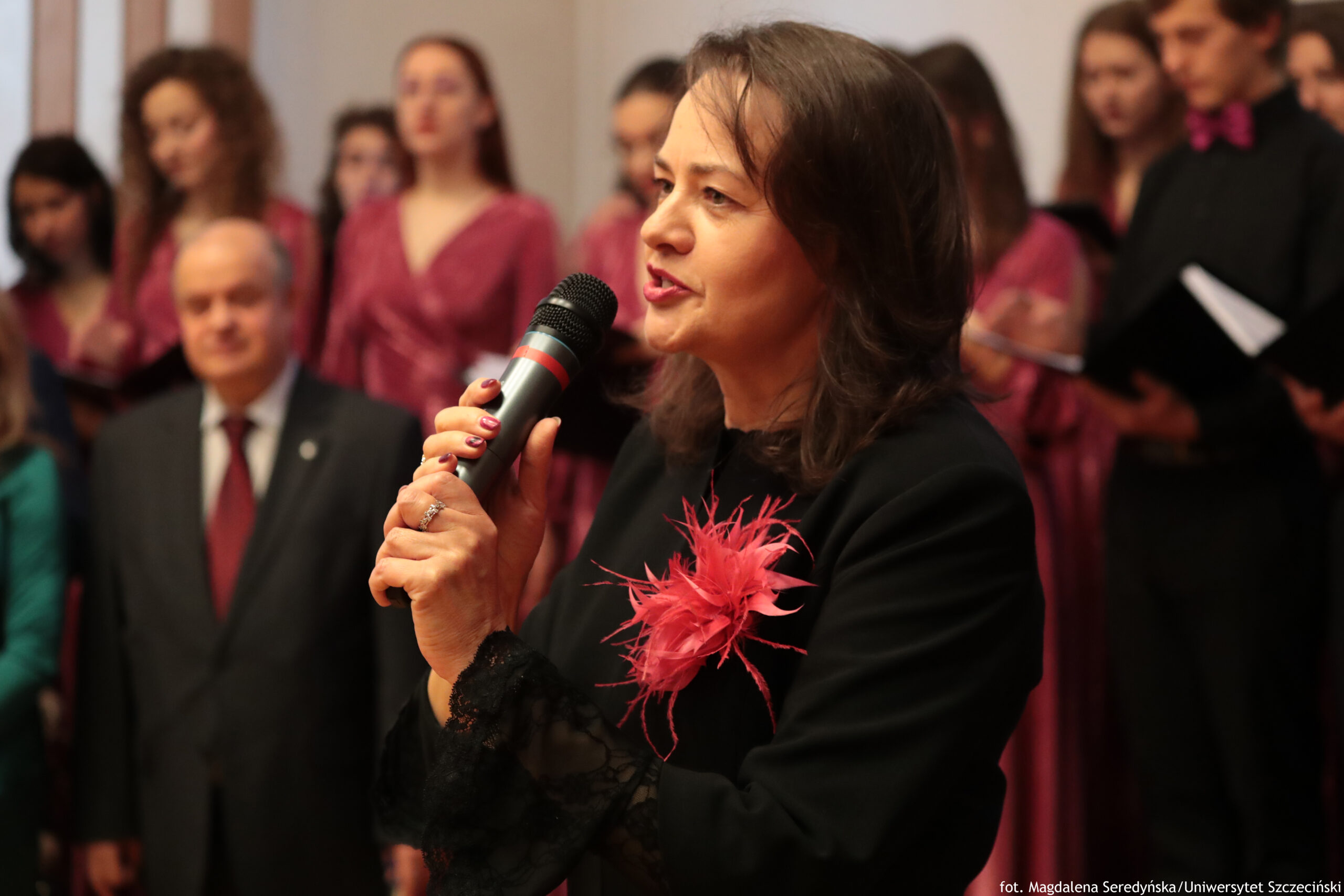 Kobieta przemawiająca do mikrofonu na tle grupy osób w strojach w odcieniach bordo. Ma jasną skórę, ciemne brązowe włosy do ramion i jest pokazana z profilu. Ubrana jest na czarno, z dużą różowo-czerwoną ozdobą z piór na piersi i koronkowymi mankietami; ma pomalowane na czerwono paznokcie i pierścionek. W tle stoi kilka kobiet w podobnych bordowych sukienkach oraz mężczyzna w garniturze z bordowym krawatem; po prawej stronie widać osoby trzymające kartki lub teczki. W prawym dolnym rogu jest podpis: “fot. Magdalena Seredyńska/Uniwersytet Szczeciński”.