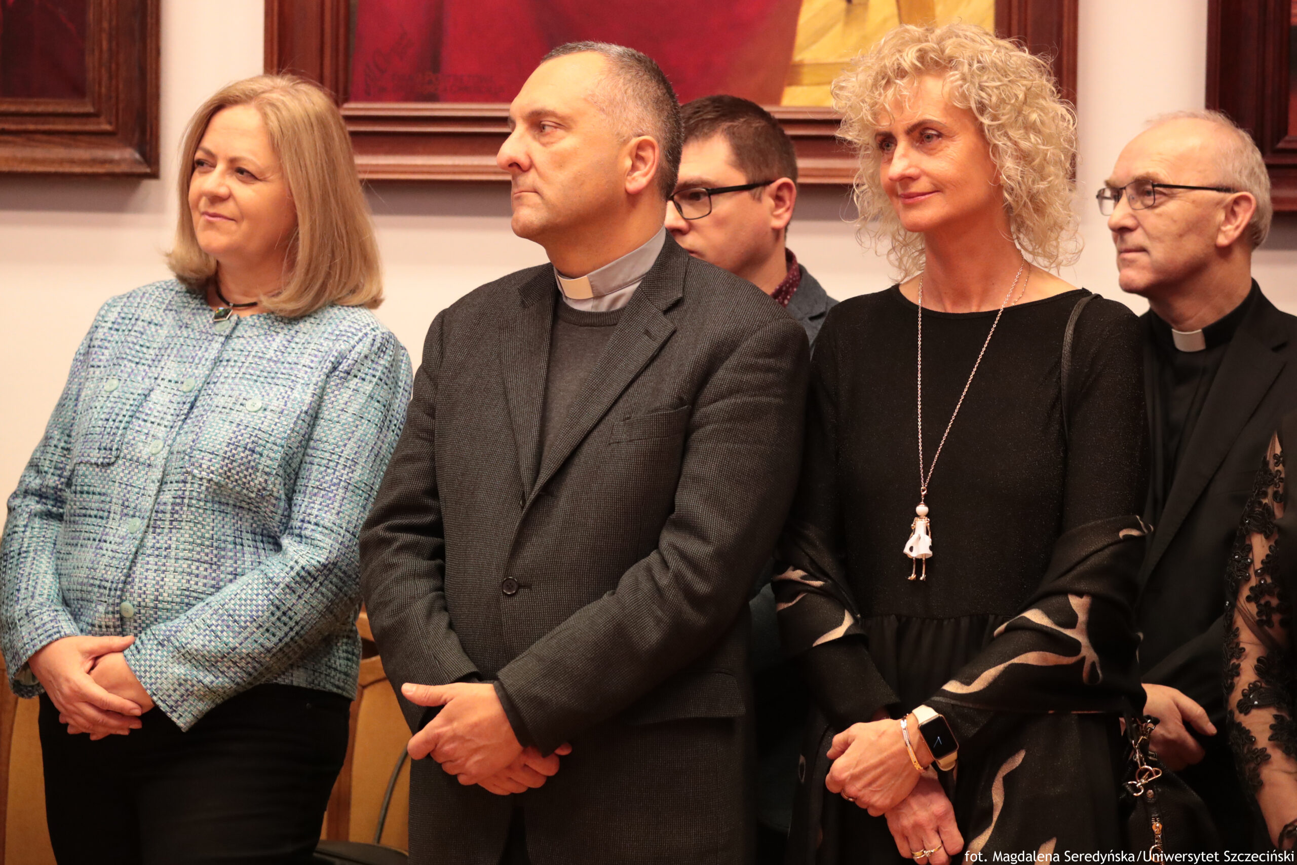Grupa pięciu dorosłych osób stojących w pomieszczeniu, sfotografowana od pasa w górę. Cztery osoby są na pierwszym planie, a jedna (mężczyzna w okularach) stoi częściowo schowana między nimi w tle. Od lewej: kobieta o jasnej skórze i jasnobrązowych, prostych włosach do ramion, w jasnoniebieskim żakiecie; ma dłonie złożone z przodu i patrzy w lewo. Obok stoi mężczyzna o jasnej skórze, krótko ostrzyżony, w ciemnej marynarce i z koloratką (strój duchownego); również ma dłonie złożone i patrzy w lewo. Za nim widać mężczyznę o jasnej skórze w okularach, z krótkimi ciemnymi włosami, patrzącego w tę samą stronę. Dalej stoi kobieta o jasnej skórze z jasnymi, kręconymi włosami, ubrana na czarno, z długim naszyjnikiem; ma ręce skrzyżowane w pasie i delikatny uśmiech. Po prawej stronie jest starszy mężczyzna o jasnej skórze w okularach, również z koloratką, w ciemnym ubraniu, patrzący w lewo. W tle wiszą duże obrazy w ciemnych ramach. W prawym dolnym rogu jest mały podpis fotograficzny: „fot. Magdalena Seredyńska / Uniwersytet Szczeciński”.