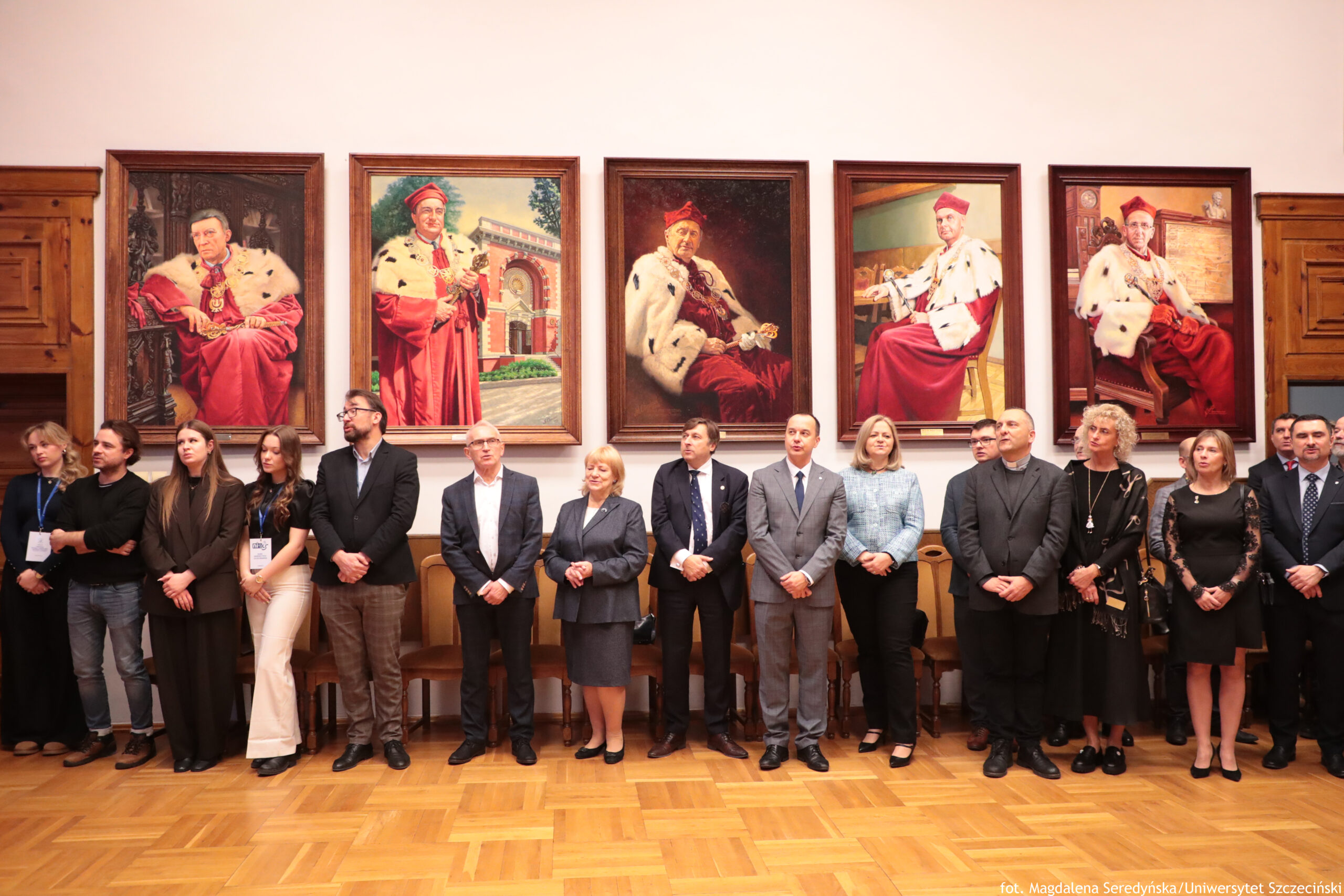 Grupa kilkunastu dorosłych osób stojących w jednym rzędzie w sali, na tle ściany z pięcioma dużymi portretami w ramach. Po portretach widać mężczyzn w czerwonych strojach ceremonialnych z białymi pelerynami z czarnymi plamami; cztery portrety pokazują postacie siedzące, jeden stojącą. Osoby w rzędzie są ubrane formalnie: garnitury, marynarki, koszule, sukienki; jedna osoba ma koloratkę i czarny strój duchowny. Pod ścianą ustawione są drewniane krzesła, a na podłodze jest parkiet. Na dole po prawej jest mały podpis: „fot. Magdalena Seredyńska/Uniwersytet Szczeciński”.