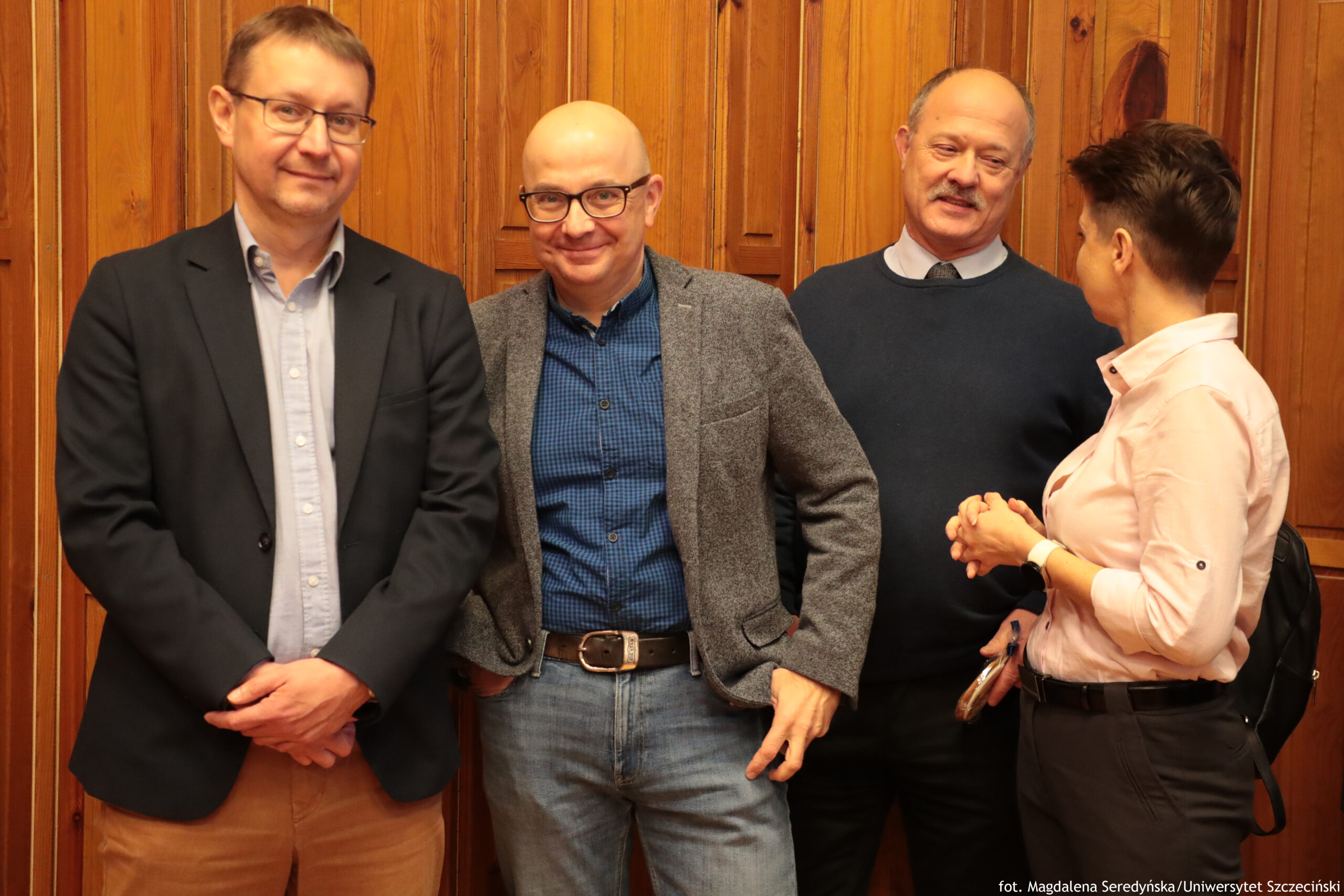 Cztery osoby stojące w pomieszczeniu na tle pionowych, drewnianych paneli. Po lewej stoi mężczyzna w okularach, w ciemnej marynarce i jasnoniebieskiej koszuli oraz beżowych spodniach; ma lekki uśmiech i dłonie złożone z przodu. Na środku stoi łysy mężczyzna w okularach, w szarej marynarce, granatowej koszuli w drobną kratkę i dżinsach; uśmiecha się, jedna dłoń jest w kieszeni. Za nim, nieco z prawej, stoi starszy mężczyzna z wąsami, w granatowym swetrze na koszuli i krawacie; uśmiecha się. Po prawej stoi osoba o krótkich, ciemnych włosach, bokiem do aparatu, w jasnoróżowej koszuli i ciemnych spodniach; ma zapięty pasek i na plecach ciemny plecak. W prawym dolnym rogu widoczny jest napis: “fot. Magdalena Seredyńska/Uniwersytet Szczeciński”.