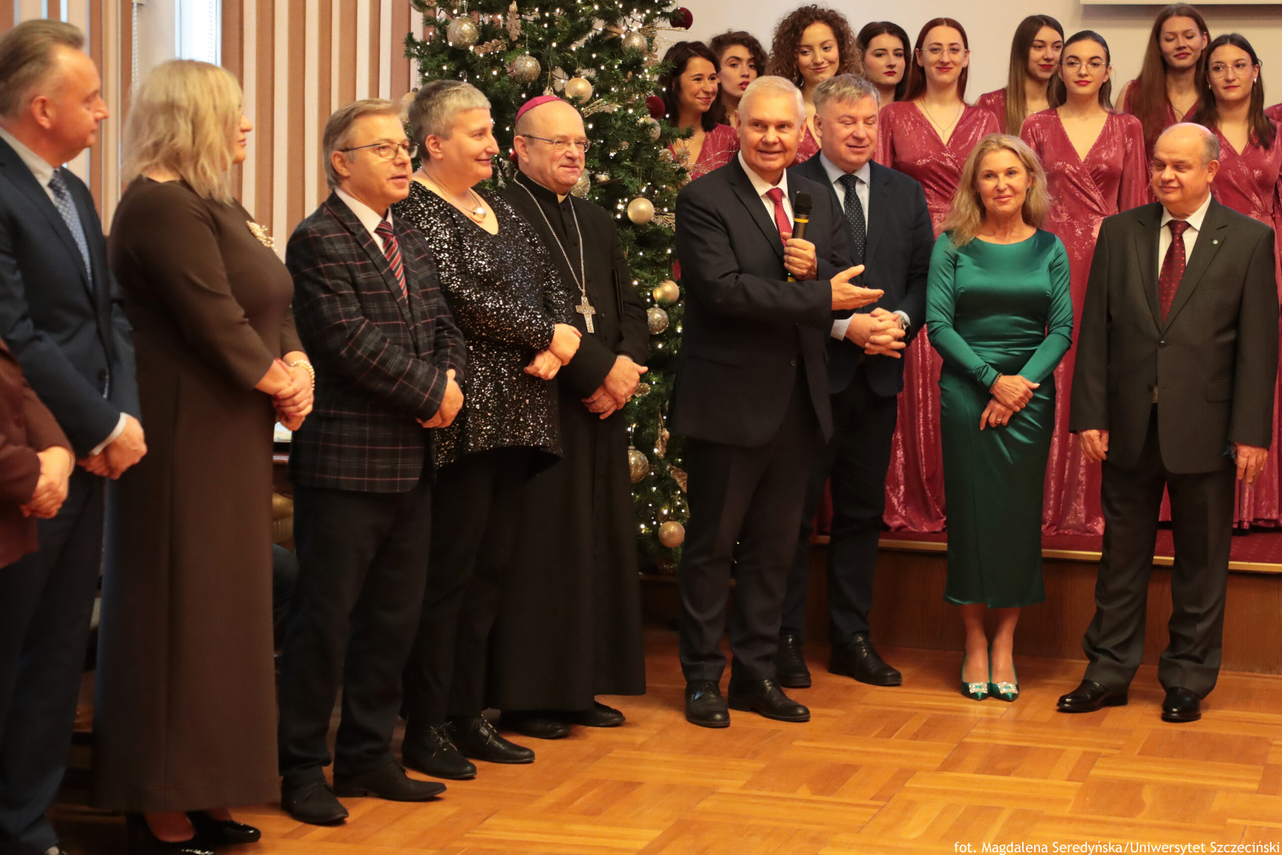 Grupa ludzi stojących w sali podczas uroczystości, z choinką bożonarodzeniową w tle. Na pierwszym planie stoi kilka osób, głównie w formalnych strojach. Pośrodku mężczyzna w ciemnym garniturze i czerwonym krawacie trzyma mikrofon i gestem dłoni wskazuje na mężczyznę stojącego obok. Obok nich stoi kobieta w zielonej, dopasowanej sukni oraz starszy mężczyzna w garniturze z czerwonym krawatem. Z lewej strony widoczna jest m.in. osoba duchowna w czarnym stroju z różową piuską i dużym krzyżem na łańcuchu, a także kobieta w czarnej, błyszczącej sukni. W tle, na podwyższeniu, stoi chór w jednakowych, połyskujących, ciemnoróżowych sukniach. Choinka jest udekorowana dużymi, jasnymi bombkami i światełkami. Na dole po prawej jest podpis: „fot. Magdalena Seredyńska/Uniwersytet Szczeciński”.