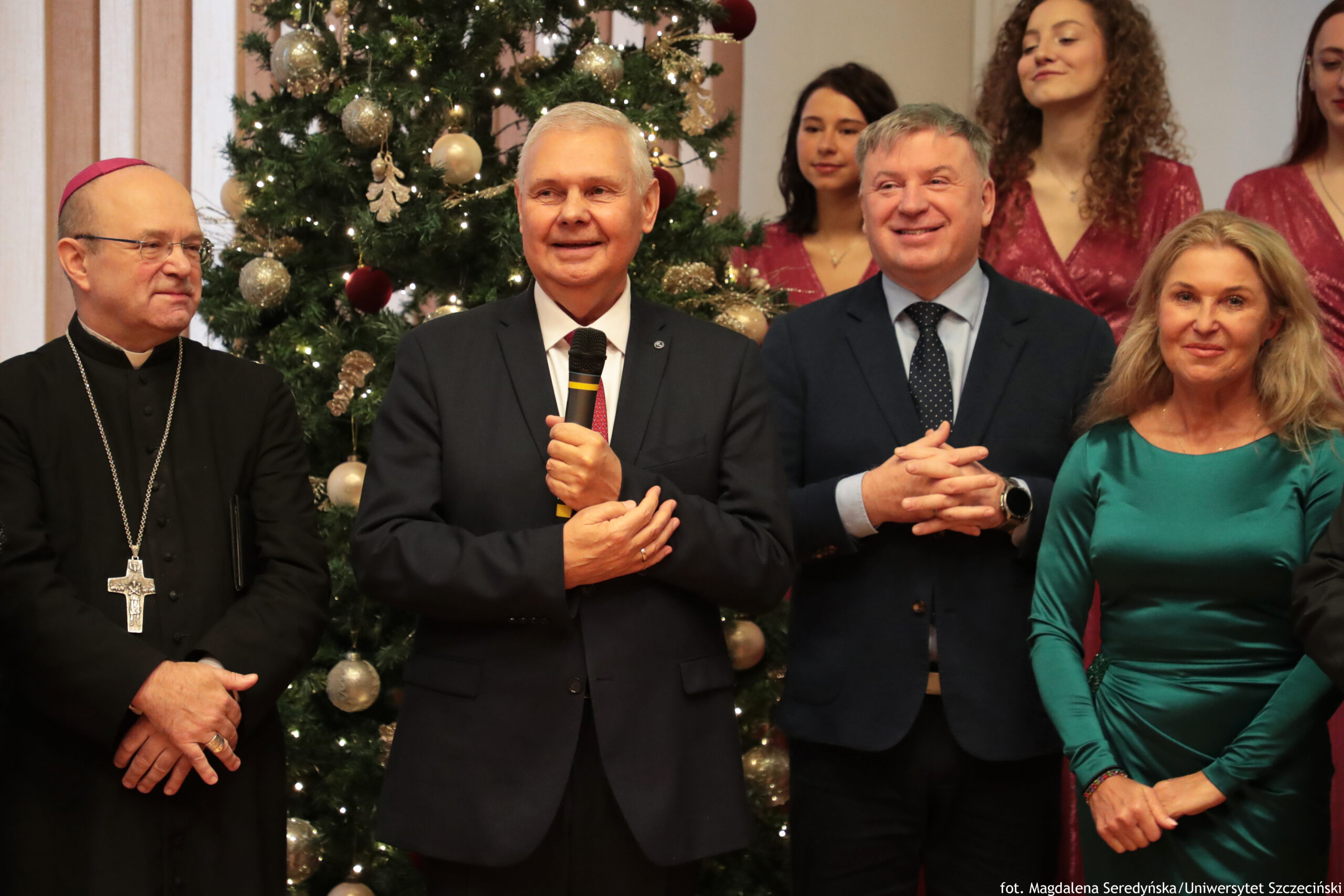 Grupa osób stojących przed udekorowaną choinką w pomieszczeniu. Na pierwszym planie widać cztery osoby: pośrodku starszy mężczyzna w ciemnym garniturze z czerwonym krawatem trzyma mikrofon i uśmiecha się; po jego lewej stronie stoi duchowny w czarnym stroju z różową piuską na głowie i dużym srebrnym krzyżem na łańcuchu; po prawej stronie jest mężczyzna w granatowym garniturze i krawacie w kropki, z dłoniami złożonymi na wysokości brzucha; skrajnie po prawej stoi kobieta w dopasowanej, błyszczącej zielonej sukni z długimi rękawami. W tle widać kilka młodych kobiet w czerwonych, błyszczących strojach. Choinka jest ozdobiona jasnymi lampkami oraz bombkami w kolorach złotym, srebrnym i bordowym. W prawym dolnym rogu jest mały podpis: „fot. Magdalena Seredyńska/Uniwersytet Szczeciński”.