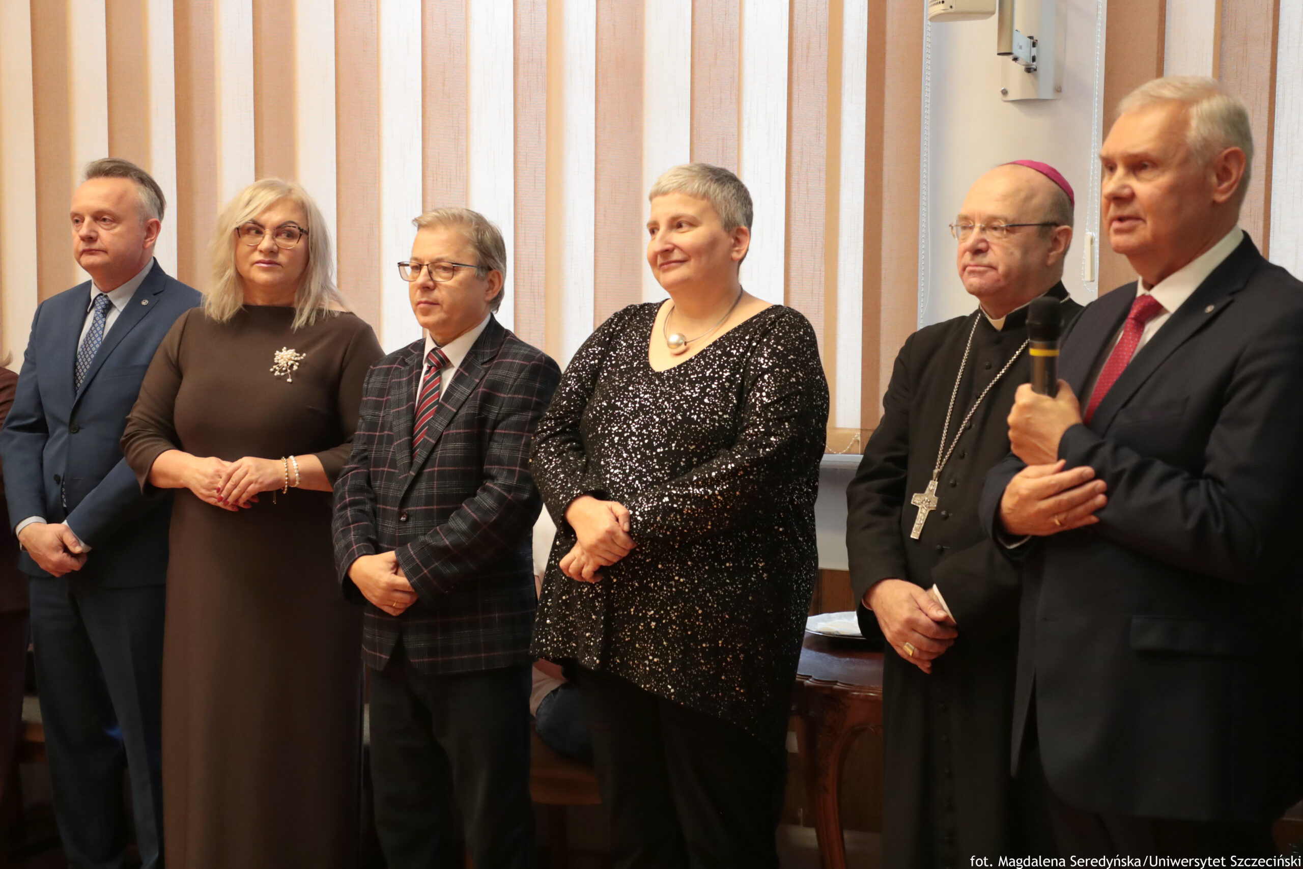 Grupa sześciu osób stojących w szeregu w pomieszczeniu, przy jasnych pionowych żaluzjach w oknie. Wszyscy są dorośli, o jasnej karnacji, ubrani elegancko. Od lewej: mężczyzna w niebieskim garniturze i krawacie; kobieta z jasnymi blond włosami i okularami w długiej brązowej sukience z ozdobą na piersi; mężczyzna w okularach w kraciastej marynarce i krawacie; kobieta z krótkimi siwymi włosami w czarnej, błyszczącej bluzce; duchowny w czarnym stroju z dużym krzyżem na łańcuchu i różową piuską; mężczyzna w ciemnym garniturze z czerwonym krawatem trzyma mikrofon, jakby przemawiał. W prawym dolnym rogu jest podpis: „fot. Magdalena Seredyńska/Uniwersytet Szczeciński”.