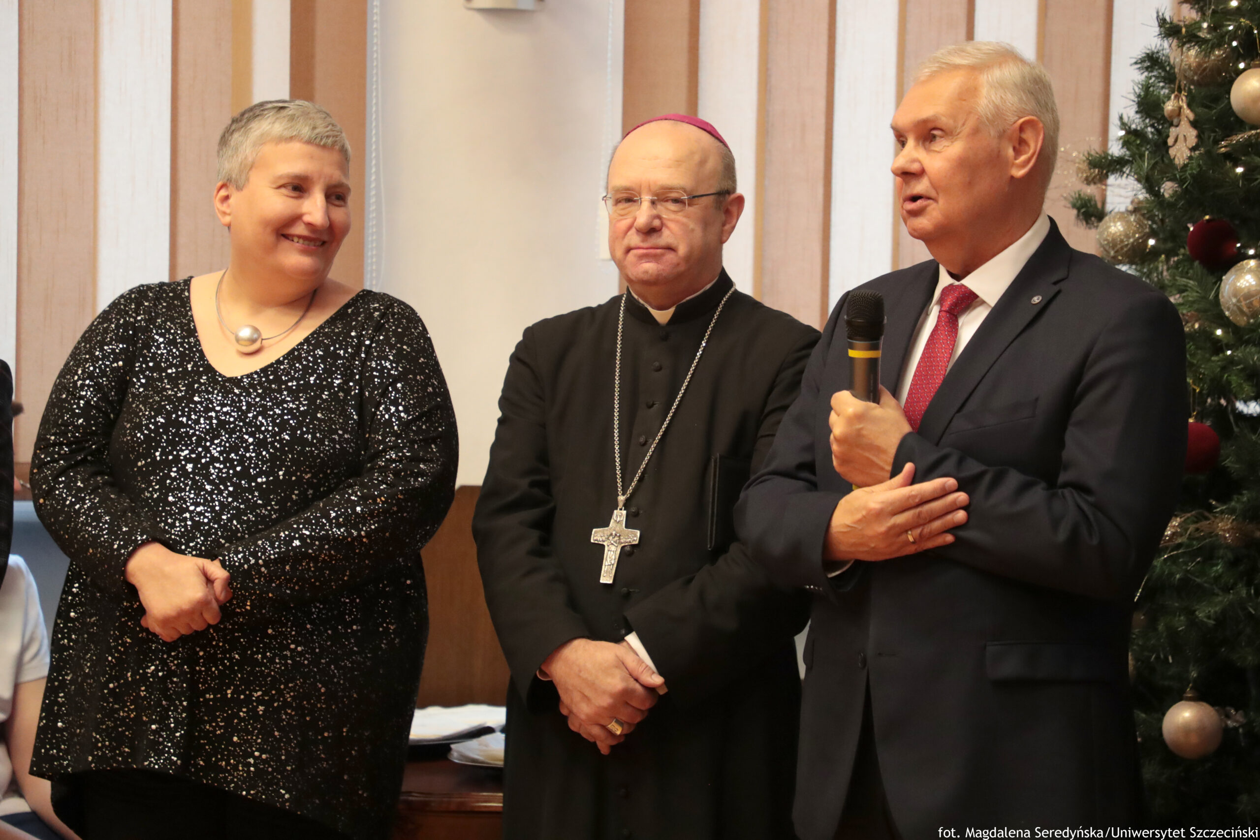 Trzy osoby stojące obok siebie w pomieszczeniu, przy choince udekorowanej bombkami. Po lewej stronie stoi uśmiechnięta kobieta o jasnej karnacji i krótkich, siwych włosach, w czarnej, błyszczącej sukience oraz z okrągłym naszyjnikiem. W środku stoi duchowny o jasnej karnacji w czarnej sutannie, z różową piuską na głowie, w okularach, z dużym krzyżem na łańcuchu; dłonie są złożone z przodu. Po prawej stronie stoi mężczyzna o jasnej karnacji w granatowym garniturze, białej koszuli i czerwonym krawacie; trzyma mikrofon i wygląda jakby przemawiał. W dolnym prawym rogu jest podpis: „fot. Magdalena Seredyńska/Uniwersytet Szczeciński”.