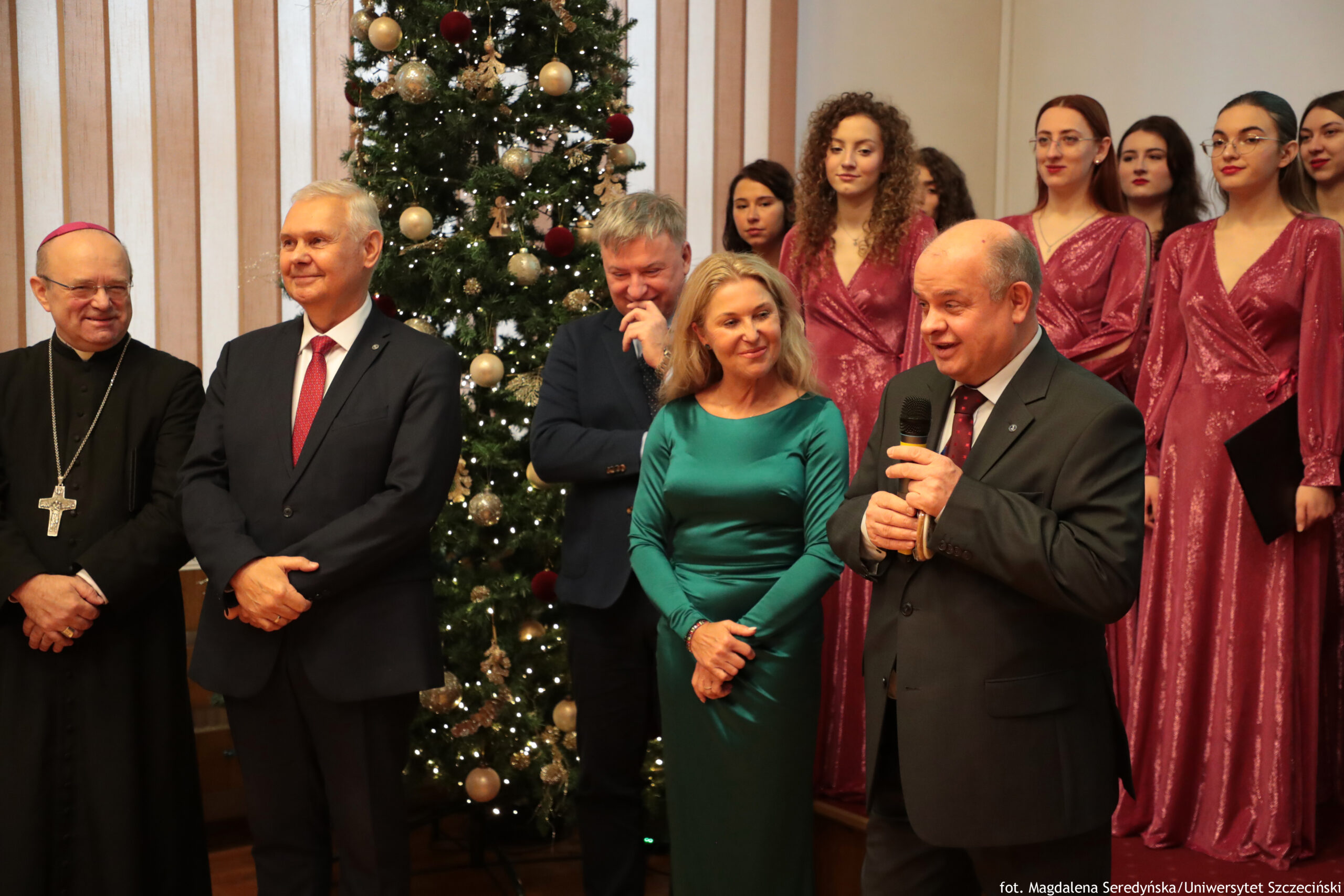 Grupa ludzi podczas uroczystości na tle dużej, udekorowanej choinki. Widać wnętrze sali i kilka osób stojących w pierwszym planie. Po lewej stronie stoi starszy mężczyzna w czarnym stroju duchownym i dużym krzyżem na łańcuchu. Obok niego stoi siwowłosy mężczyzna w ciemnym garniturze z czerwonym krawatem. Za nimi, częściowo zasłonięty, stoi mężczyzna w granatowym garniturze, z ręką przy twarzy. Na środku stoi kobieta w zielonej, satynowej sukni z długim rękawem. Po prawej stronie stoi łysy mężczyzna w ciemnym garniturze, trzyma mikrofon i wygląda, jakby przemawiał. W tle, po prawej stronie choinki, stoi kilka młodych kobiet w jednakowych, połyskujących bordowych sukniach; część z nich ma rozpuszczone włosy, jedna ma okulary. Choinka jest wysoka, z białymi i złotymi bombkami oraz światełkami. W prawym dolnym rogu jest podpis: „fot. Magdalena Seredyńska / Uniwersytet Szczeciński”.