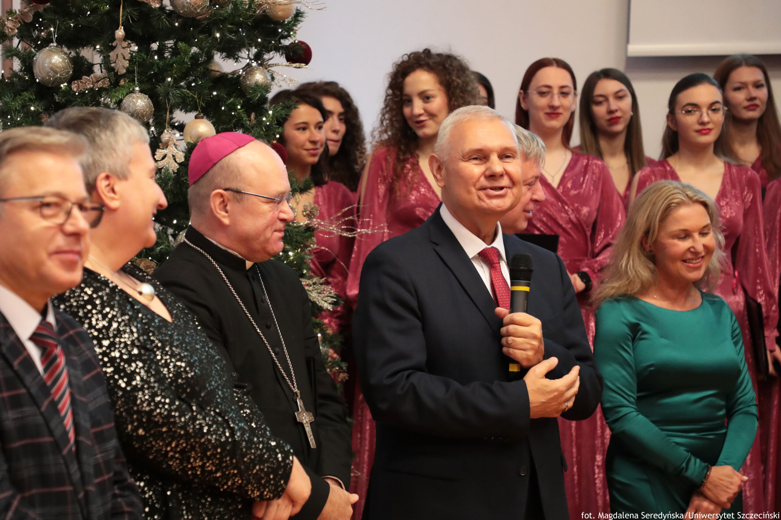Grupa ludzi podczas uroczystości świątecznej w pomieszczeniu, obok dużej choinki udekorowanej srebrnymi i złotymi bombkami oraz światełkami. W centrum stoi starszy mężczyzna o siwych włosach w ciemnym garniturze i czerwonym krawacie, trzyma mikrofon i przemawia z uśmiechem. Po jego lewej stronie stoi duchowny w czarnej sutannie z łańcuchem i krzyżem oraz fioletową piuską; obok niego widać kobietę w czarnej, błyszczącej sukni. Po prawej stronie stoi kobieta w zielonej sukience, uśmiechnięta, z długimi blond włosami. W tle znajduje się kilka młodych kobiet w bordowych, połyskujących strojach, ustawionych w rzędzie jak chór. W prawym dolnym rogu jest mały podpis: „fot. Magdalena Seredyńska/Uniwersytet Szczeciński”.