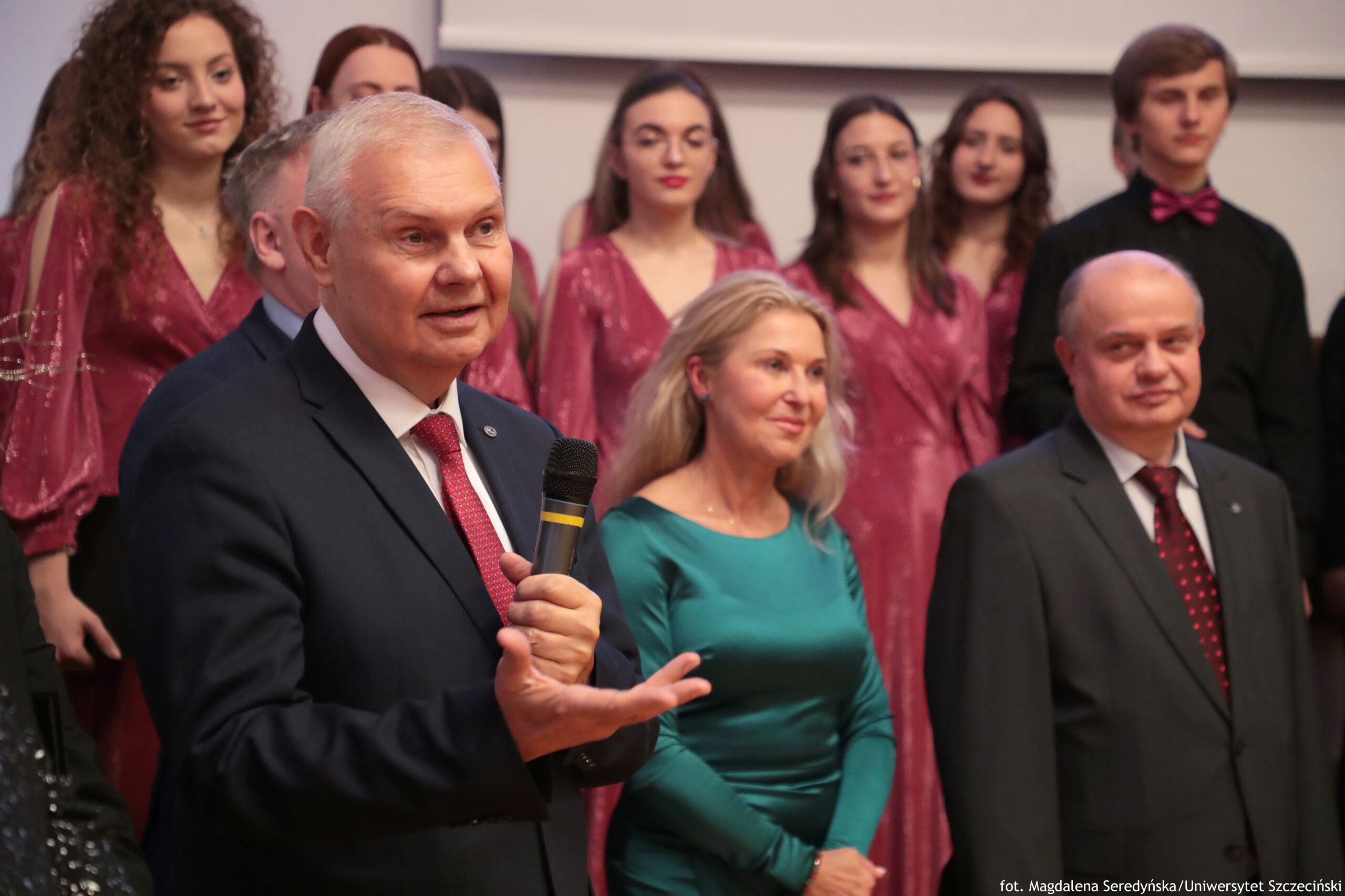 Starszy mężczyzna w granatowym garniturze i czerwonym krawacie, o krótkich siwych włosach i jasnej skórze, przemawiający do mikrofonu w pomieszczeniu. W kadrze na pierwszym planie mężczyzna trzyma mikrofon i gestykuluje dłonią, jak podczas wystąpienia. Obok niego stoi kobieta o jasnej skórze i długich blond włosach, ubrana w zieloną sukienkę. Po prawej stronie stoi łysy mężczyzna o jasnej skórze w ciemnym garniturze i czerwonym krawacie w drobne wzory. W tle widać grupę młodych osób, głównie kobiet o jasnej skórze, ubranych w jednakowe bordowe stroje, oraz młodego mężczyznę w czarnym stroju z bordową muszką. Na dole po prawej znajduje się mały napis: „fot. Magdalena Seredyńska/Uniwersytet Szczeciński”.